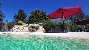 Una piscina cristalina con una sombrilla roja, una cascada y un muro de piedra.