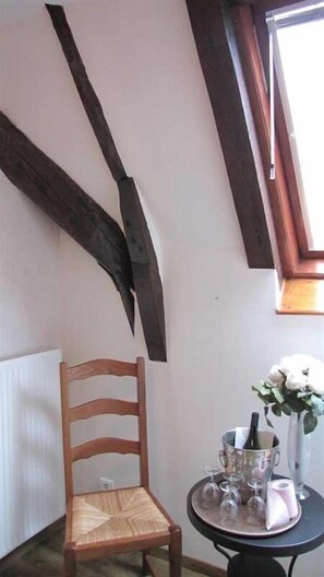 Une pièce avec une chaise en bois, une petite table ornée d’un vase et de verres, et un plafond en pente avec des poutres apparentes.