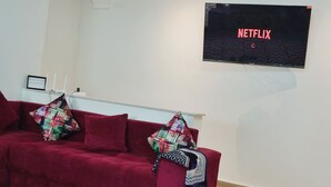 Ein rotes Sofa mit gemusterten Kissen, ein rot-weißer Teppich und ein an der Wand befestigter Fernseher, der 'NETFLIX' anzeigt, im Wohnzimmer.