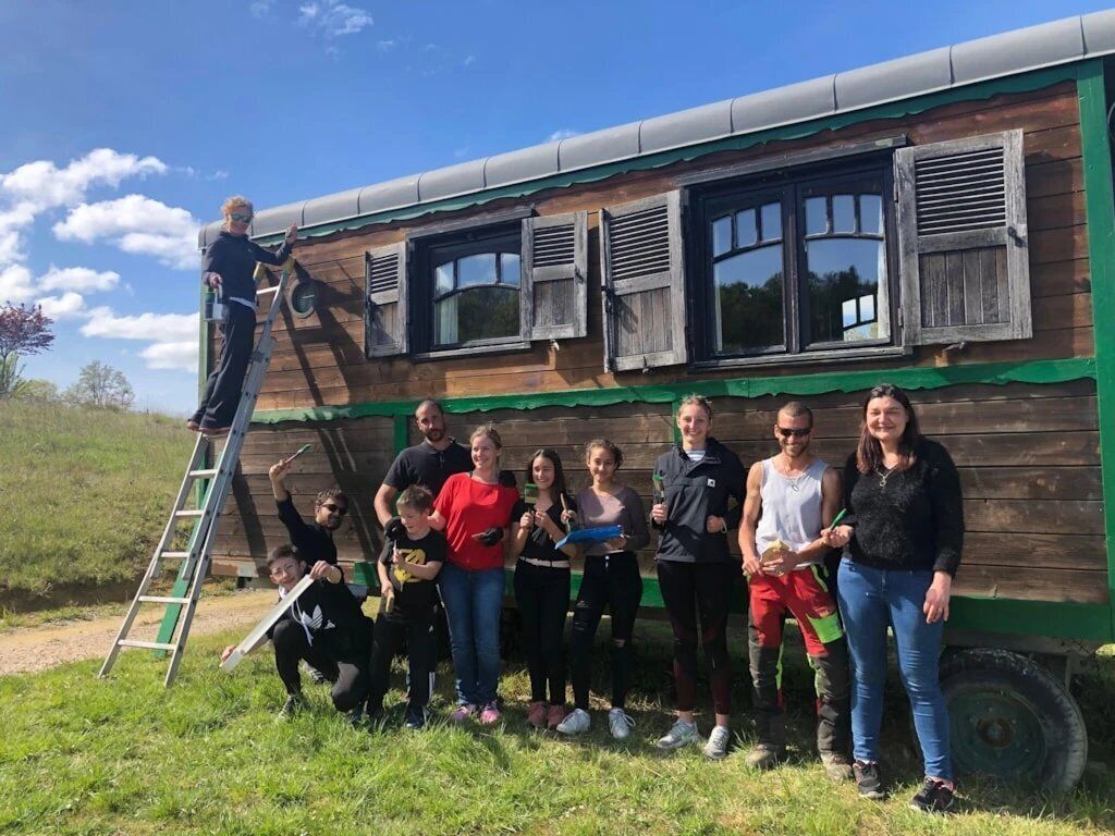 Un grupo de personas frente a una construcción de madera con una escalera y un vehículo.