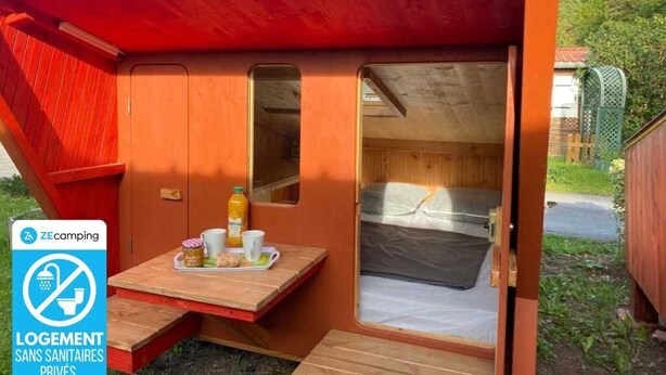 A small, single-room cabin with a wooden exterior, a door leading to a sleeping area, and a small table with breakfast items. - ZEcamping 2* dog friendly locatif 2 pers (Angicourt)