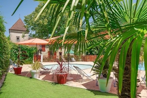 Area piscina con ombrellone rosso, piante in vaso e un edificio con tetto rosso sullo sfondo.