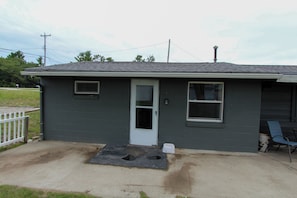 A small single-story building with a dark gray exterior, a white door, and two windows. There is a concrete patio with a metal step leading to the door. - Escape to your private Lake Huron retreat with 6 fully equipped cottages, 13 bedrooms, and 6 bathrooms, perfect for family reunions, weddings, retreats, and group getaways. (Iosco County)
