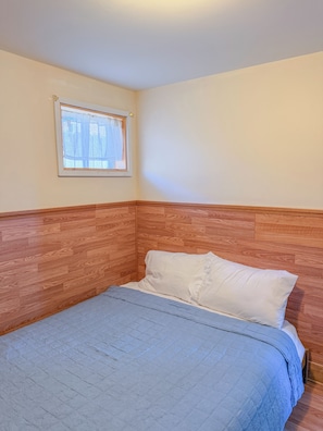 Ein Einzelbett mit Holz-Kopfstück, ein Fenster mit leichtem Vorhang und eine schlichte Wand.