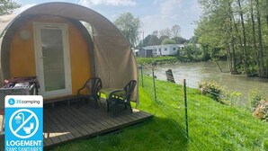 Een kampeertent met een deur en een klein terras, omgeven door een grasveld en een waterpartij.