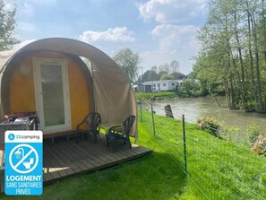 Ein Campingzelt mit Tür und einer kleinen Holzterrasse, umgeben von einer Wiese und einem Gewässer.