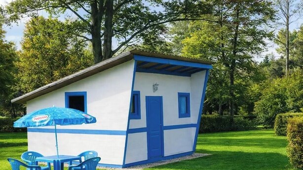 A small, single-story building with a blue door and windows, a table with blue chairs, and a blue umbrella.