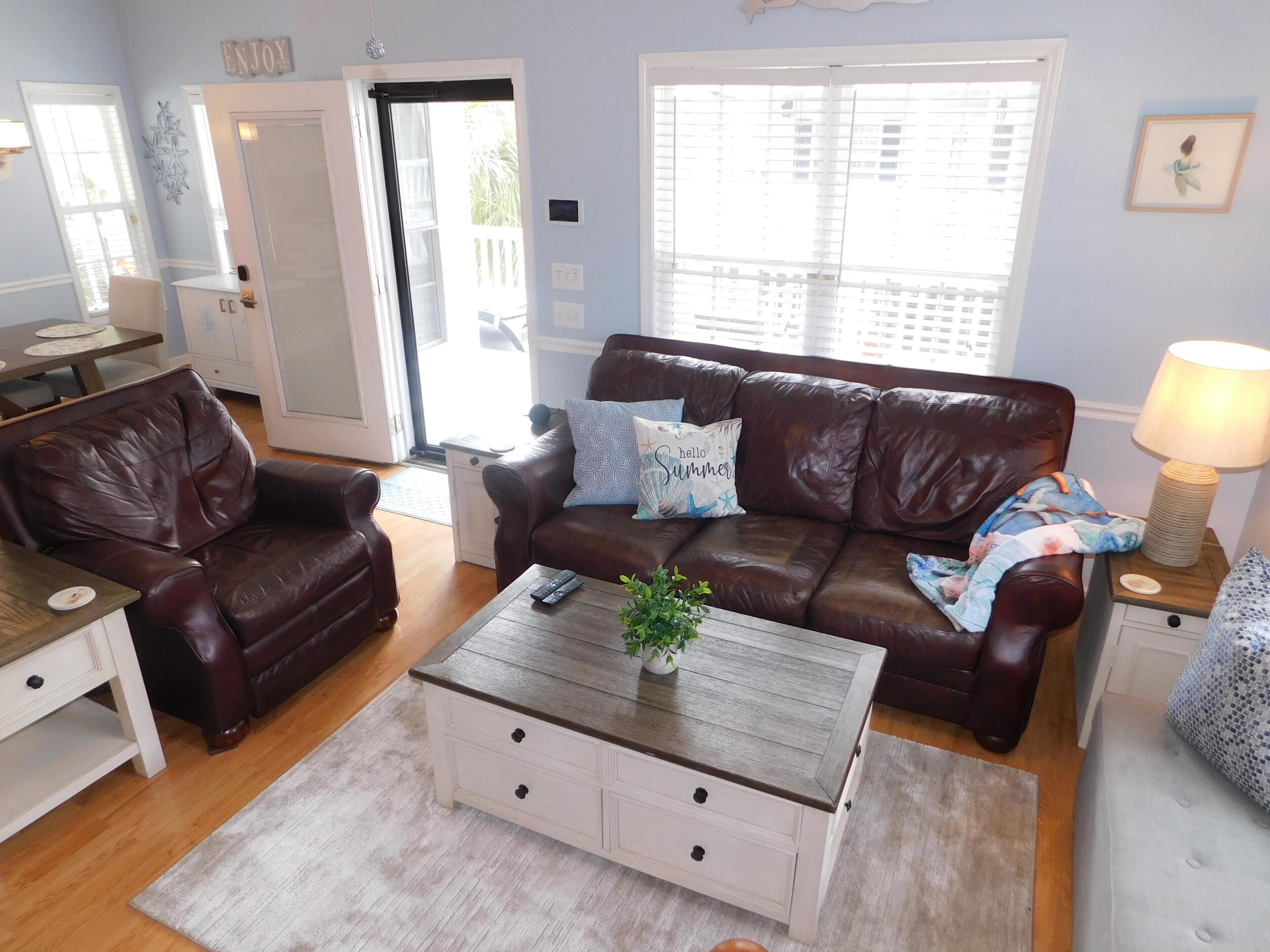 A living room with a brown leather sofa, a white coffee table with drawers, a wooden side table with a lamp, and a view of the outside through a window.