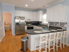 A modern kitchen with white cabinetry, stainless steel appliances, a central island with bar stools, and a trash can. - Ocean Lakes Site 1705 "A Mermaids Treasure" 4bed/3bath | golf cart (Myrtle Beach)
