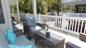 A covered outdoor seating area with wicker furniture, a white fence, and palm trees in the background. - Ocean Lakes Site 1705 "A Mermaids Treasure" 4bed/3bath | golf cart (Myrtle Beach)