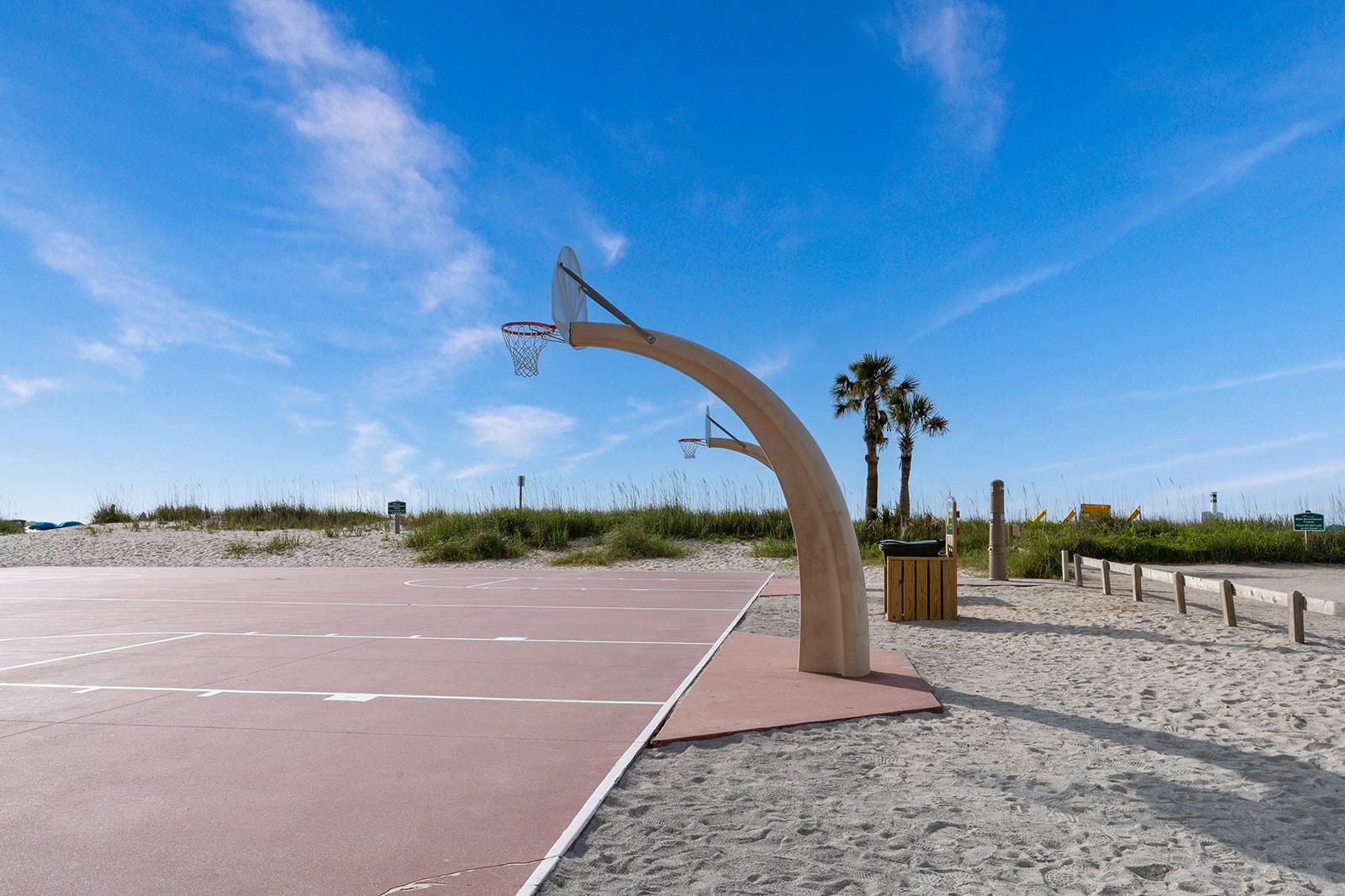 An outdoor basketball court with a unique curved hoop structure, surrounded by sand and palm trees.