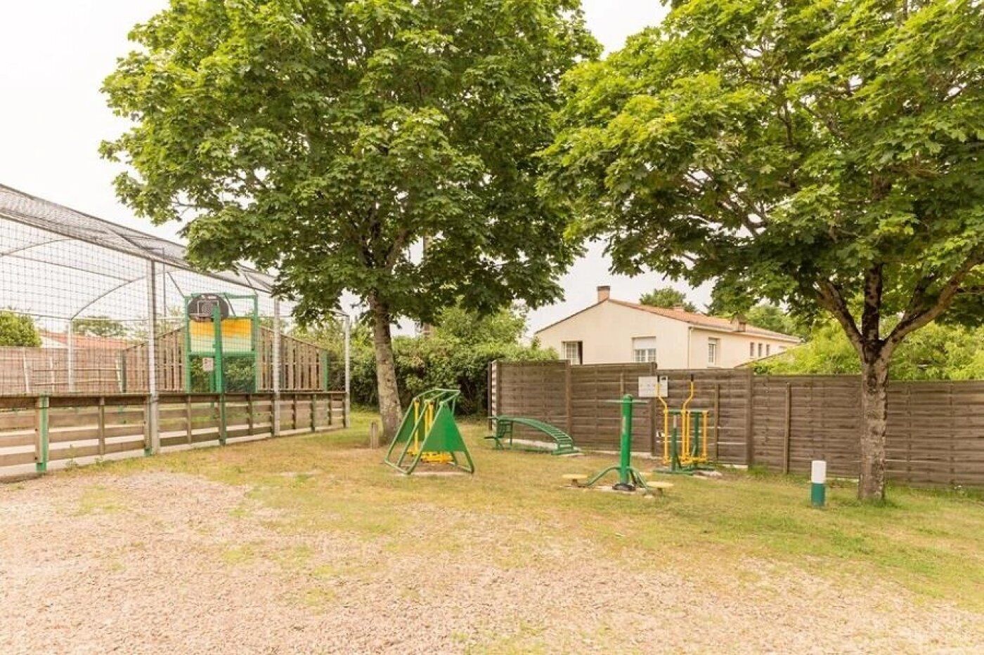 A fenced outdoor area with playground equipment, trees, and a building in the background.