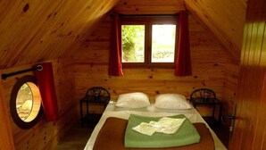 Une chambre dans une cabane en bois, avec un lit, deux chaises, une fenêtre et un miroir rond.