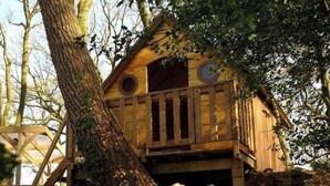 Une cabane en bois dotée d’un balcon, entourée d’arbres et d’un mur de pierres.
