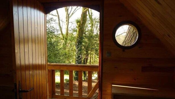 Une cabane en bois dotée d’une terrasse, d’une fenêtre ronde et d’une porte donnant sur un espace extérieur.