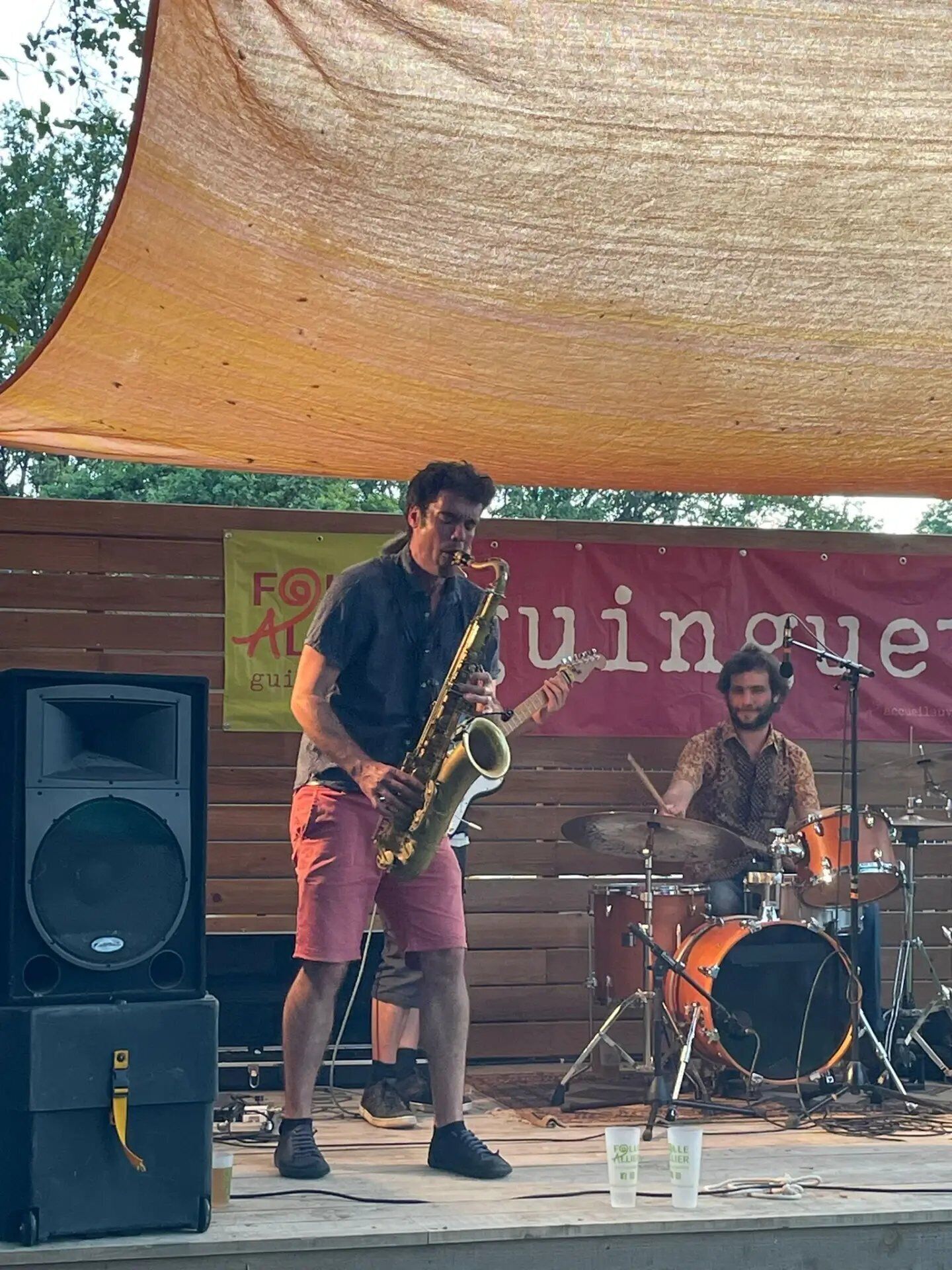 Un músic tocant un saxofon a un escenari exterior, amb una bateria de fons.