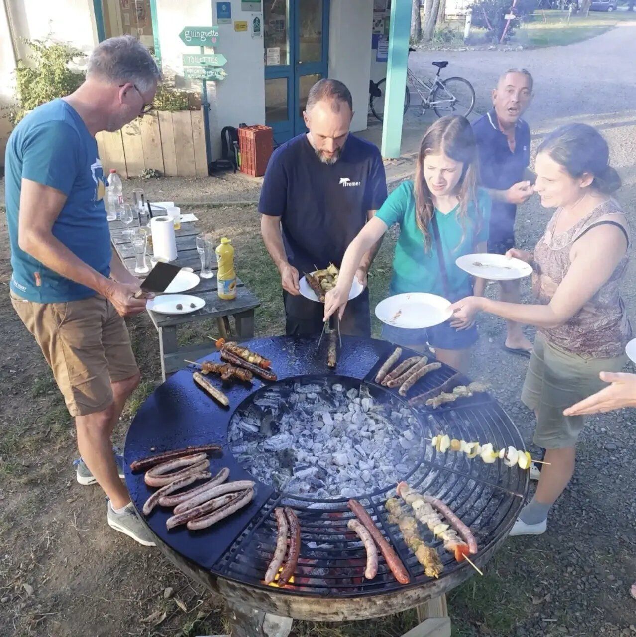A group of people around a barbecue grill with sausages and skewers being cooked.