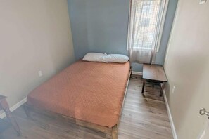 A small, single-bed room with a wooden floor, a window with sheer curtains, and a small wooden table.