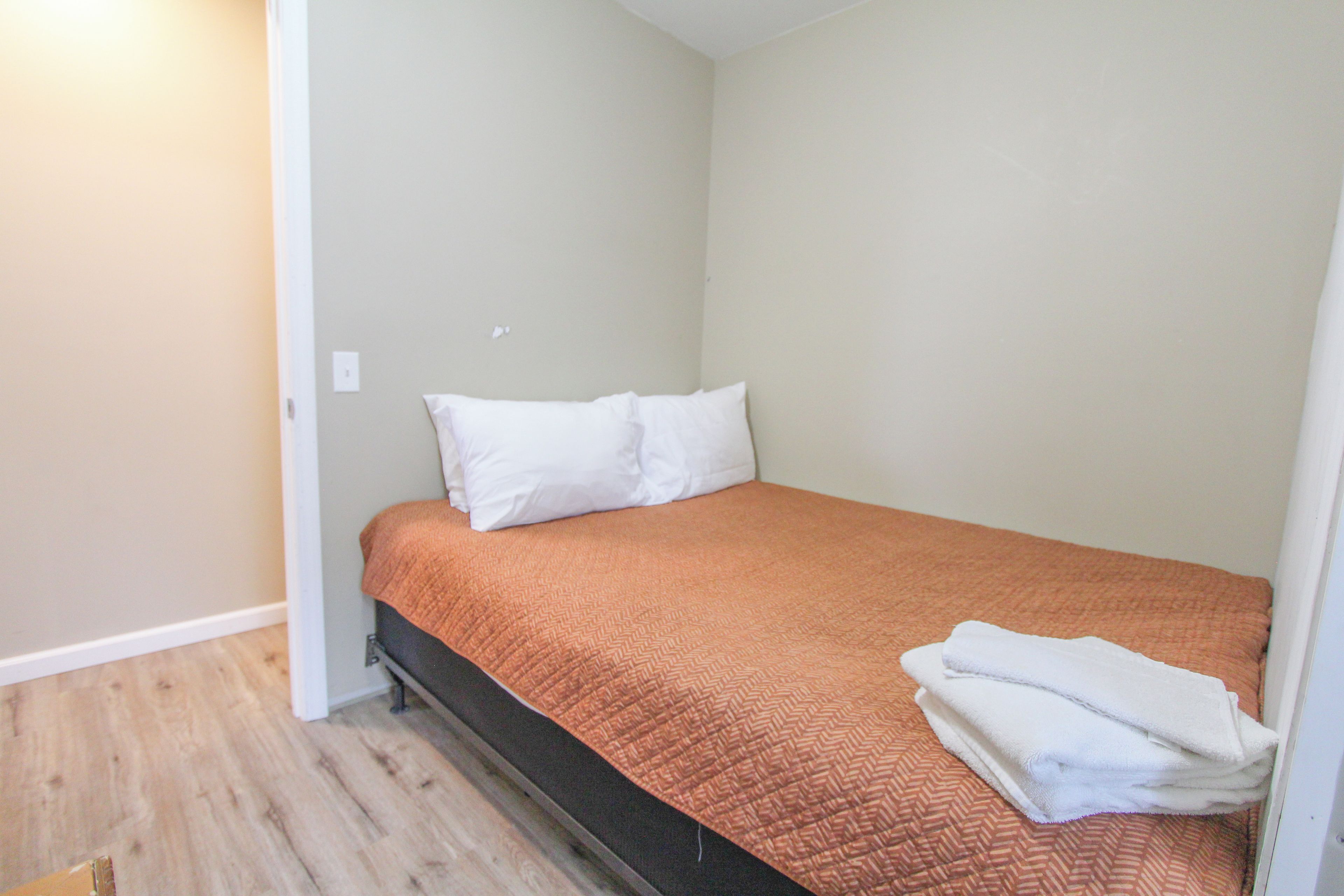 A small, single-bed room with a wooden floor, white walls, and a brown bedspread.