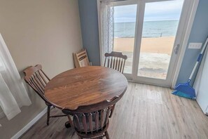 A room with a wooden dining table and chairs, a large sliding glass door leading to a beach view.