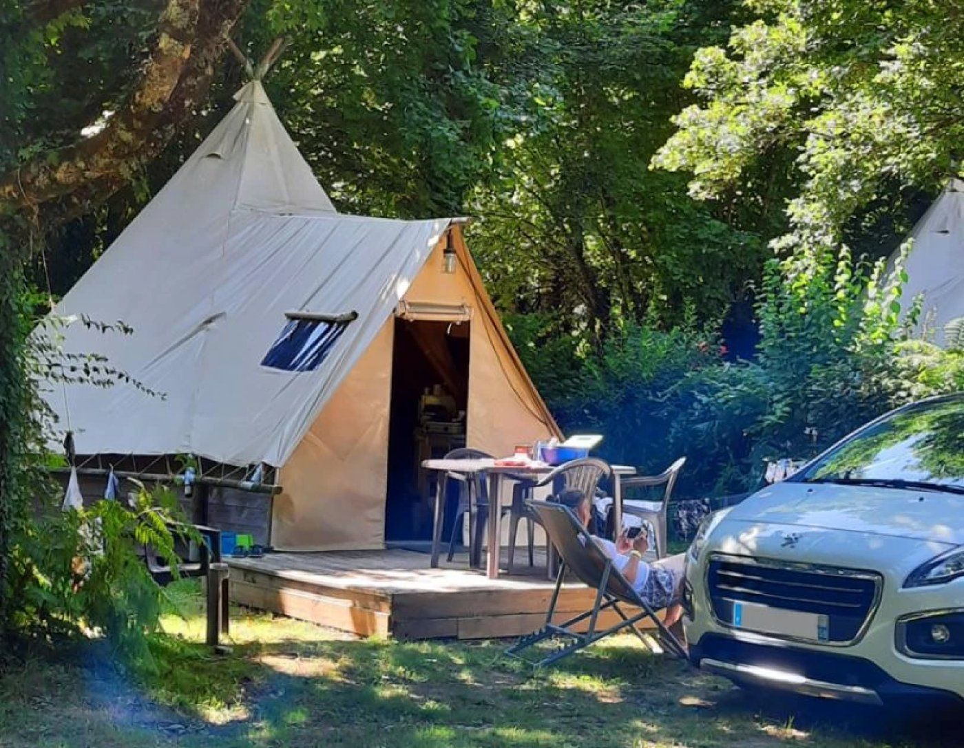 A tent with a table and chairs outside, a car parked nearby, and a person reclining on a chair.