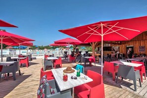 Ein Essbereich im Freien mit roten Sonnenschirmen, einer Holzdielen-Terrasse und Tischen mit Stühlen.