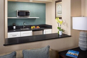 Ein kompaktes Hotelzimmer mit Küchenzeile, inklusive Mikrowelle, Spüle und einer kleinen Essecke. Auf dem Nachttisch befinden sich ein Blumentopf und eine Lampe.