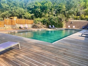 Une piscine rectangulaire avec une terrasse en bois, entourée de chaises longues et d’un mur en pierre.
