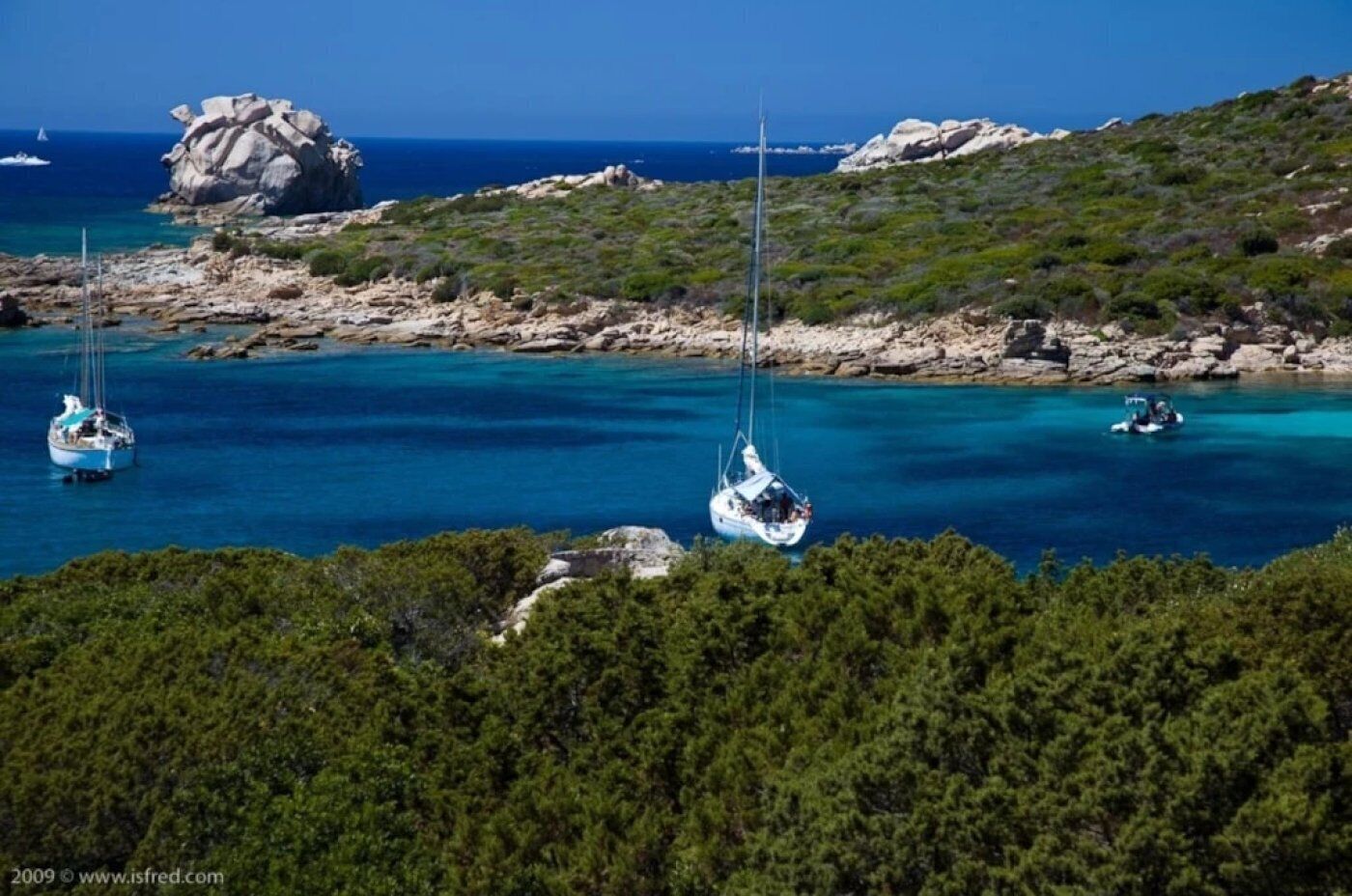 A coastal landscape with clear turquoise waters, rocky outcrops, and dense green vegetation.