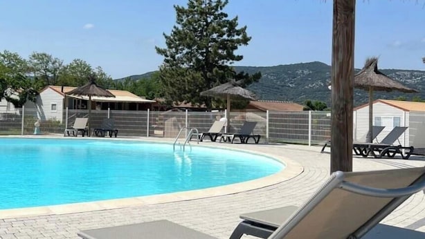 Piscina con sillas de descanso y sombrillas, rodeada por una valla y árboles.