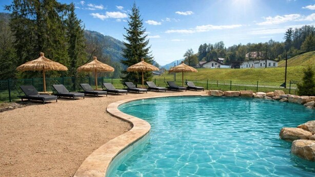 A swimming pool with lounge chairs and thatched umbrellas, surrounded by a grassy area and mountains in the background.