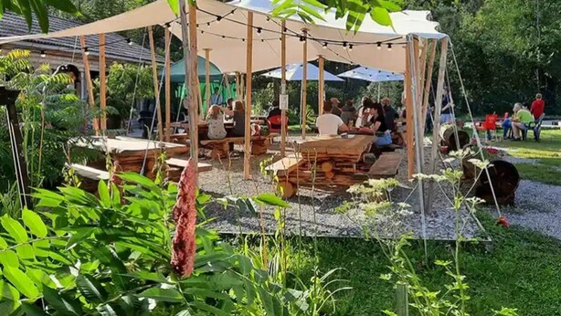 An outdoor dining area with wooden tables and benches, surrounded by greenery and a tent structure.