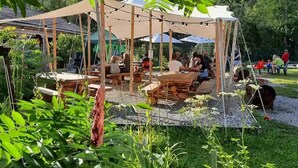 An outdoor dining area with wooden tables and benches, surrounded by greenery and a tent structure.