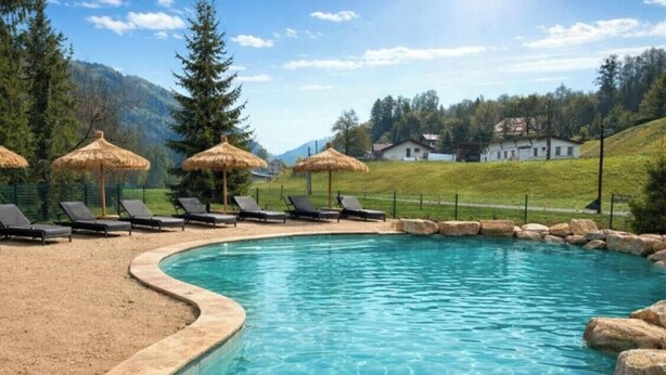 A swimming pool with lounge chairs and umbrellas, surrounded by a pebble beach and a mountainous backdrop.
