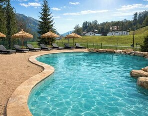 A swimming pool with lounge chairs and umbrellas, surrounded by a pebble beach and a mountainous backdrop.