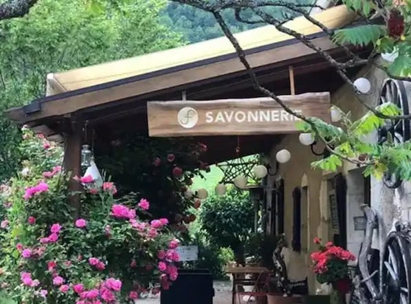 A covered outdoor seating area with a sign reading 'SAVONNERIE,' surrounded by lush greenery and flowering plants.