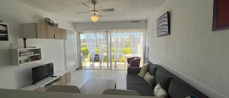 Uma sala de estar moderna com um sofá, uma TV de tela plana, um ventilador de teto e vista para uma área externa por meio de portas de vidro.