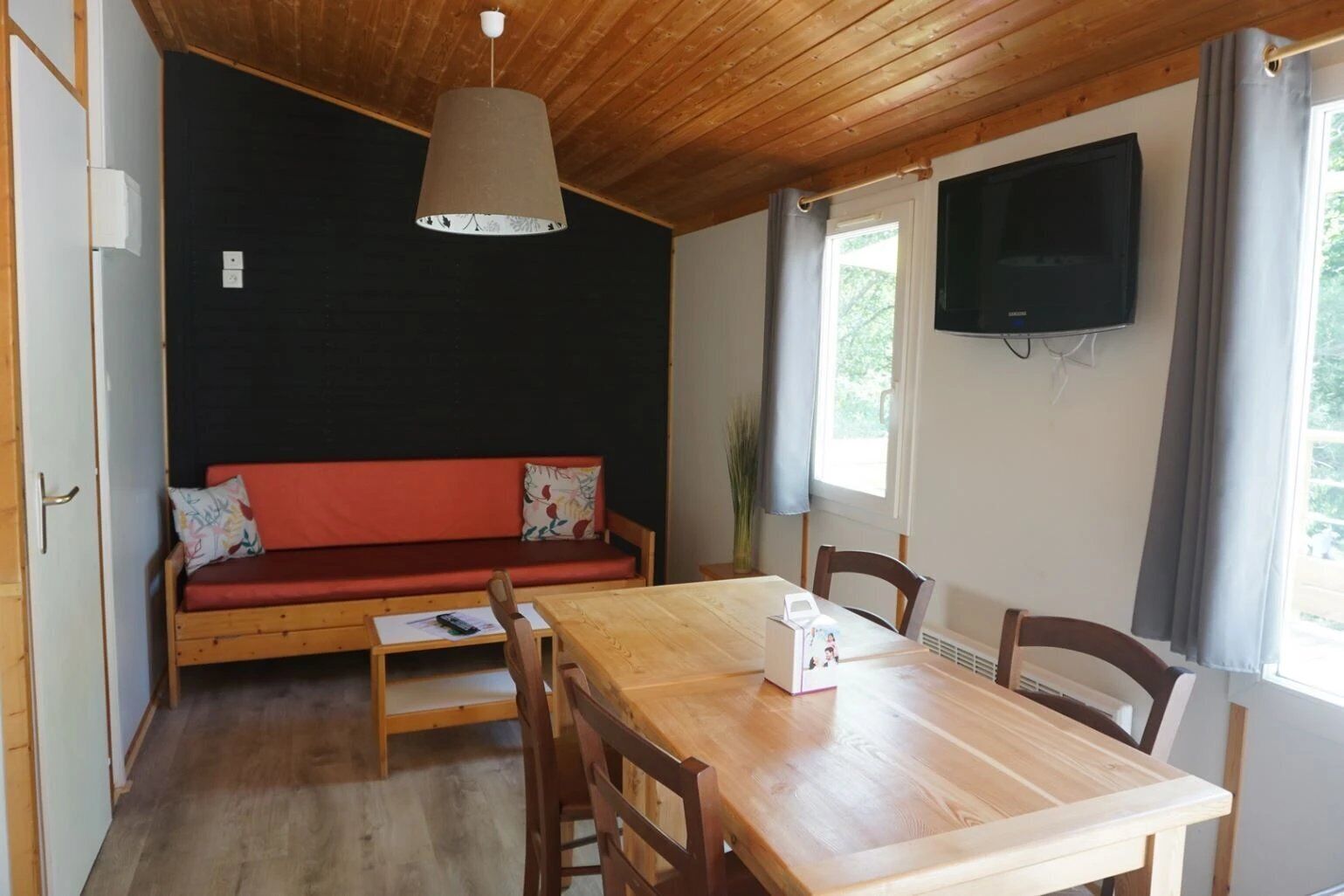A room with a wooden ceiling, a red sofa with cushions, a wooden dining table with chairs, a television on the wall, and a window with curtains.