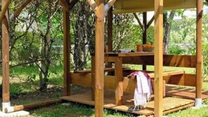 Um gazebo de madeira com uma mesa e bancos, rodeado por árvores.