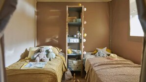 Una habitación pequeña y acogedora con dos camas, piso de madera, una estantería con objetos personales y una ventana.