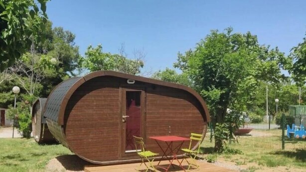 Una estructura de madera con una puerta roja, un pequeño patio con una mesa y sillas, y un área verde circundante.