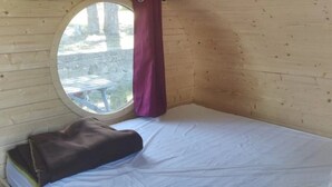 Una cama individual con colchón blanco, una manta marrón y un ventanal redondo con cortina morada.