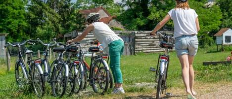 Una persona ajustando una bicicleta, otra caminando cerca, y varias bicicletas estacionadas en un área verde con árboles y una valla de madera al fondo.