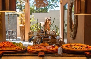 A restaurant with three pizzas on wooden boards, a dining table with chairs, and a statue in the background. - ZEcamping 3* piscine animaux acceptés locatif 5 pers (Tuchan)