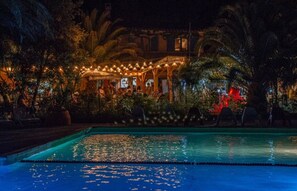 Un área de piscina nocturna con superficie de agua cristalina, rodeada de palmeras y un edificio con luces colgantes.