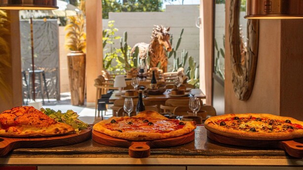 A restaurant with three pizzas on wooden boards, a dining area with tables and chairs, and a view of a garden with plants and a horse statue. - ZEcamping 3 étoiles piscine animaux acceptés tente 12m2 2 pers (Tuchan)