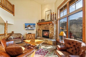 A cozy living room with leather furniture, a stone fireplace, and a large window overlooking a mountainous landscape.