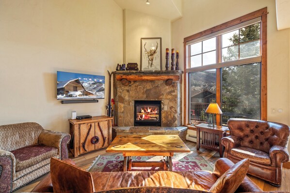 A cozy living room with a stone fireplace, leather furniture, and a large window offering a view of the outdoors.