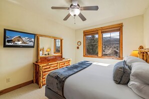A bedroom with a bed, a wooden dresser, a television mounted on the wall, a window with a view, and a ceiling fan.