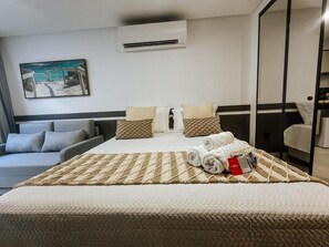 A hotel room with a bed, a sofa, an air conditioning unit, and a painting on the wall.
